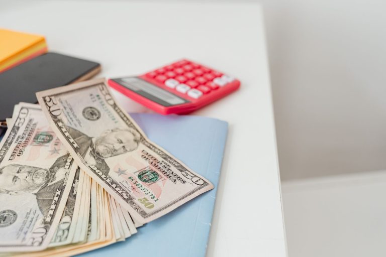 Fifty dollar bills fanned out on a desk beside a red calculator and a notebook