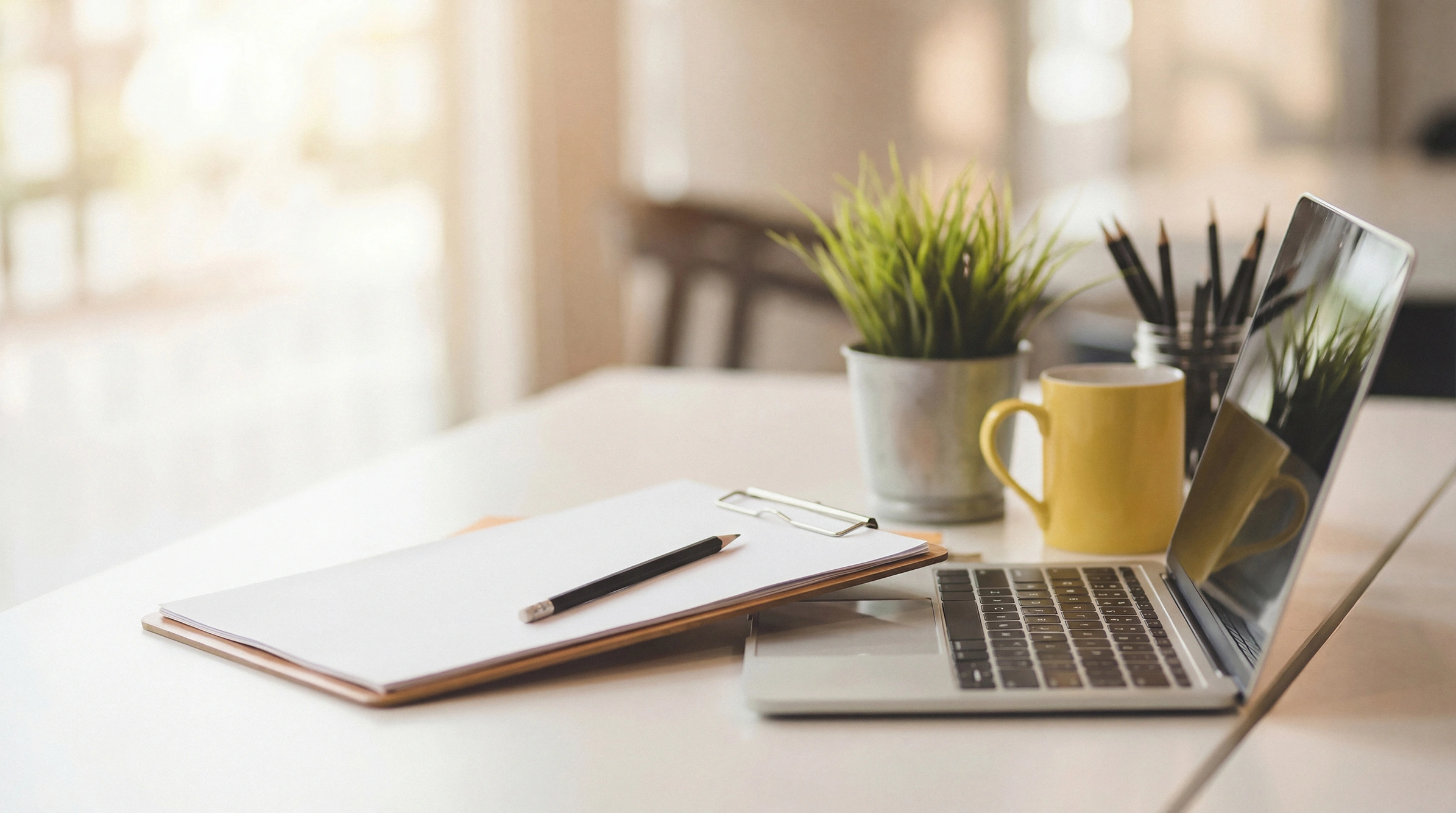 A clean, naturally lit home-office desk with a laptop, notebook, and a small plant